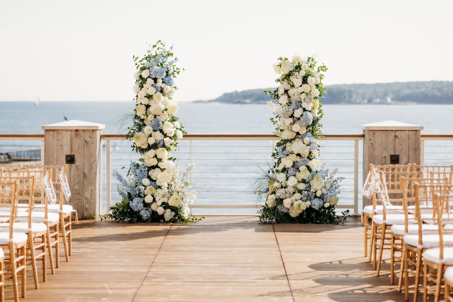 Elegant outdoor wedding ceremony setup with white floral arrangements and wooden chairs overlooking a waterfront view.