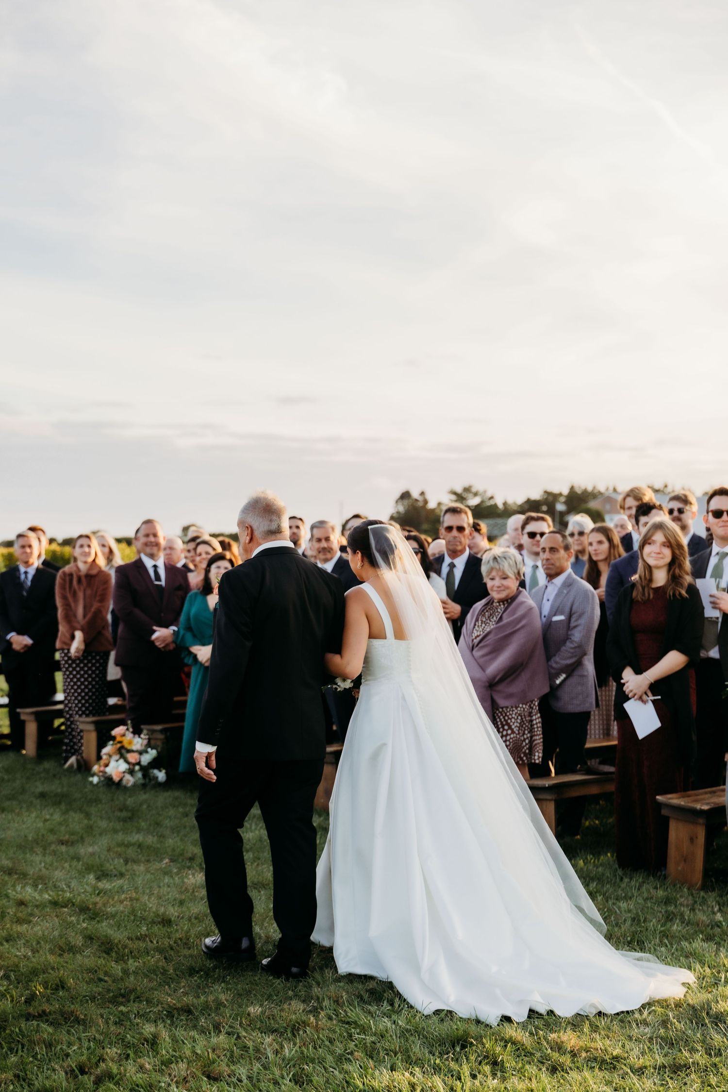 A large outdoor wedding ceremony takes place on a grassy hillside with guests gathered to witness the celebration at sunset.