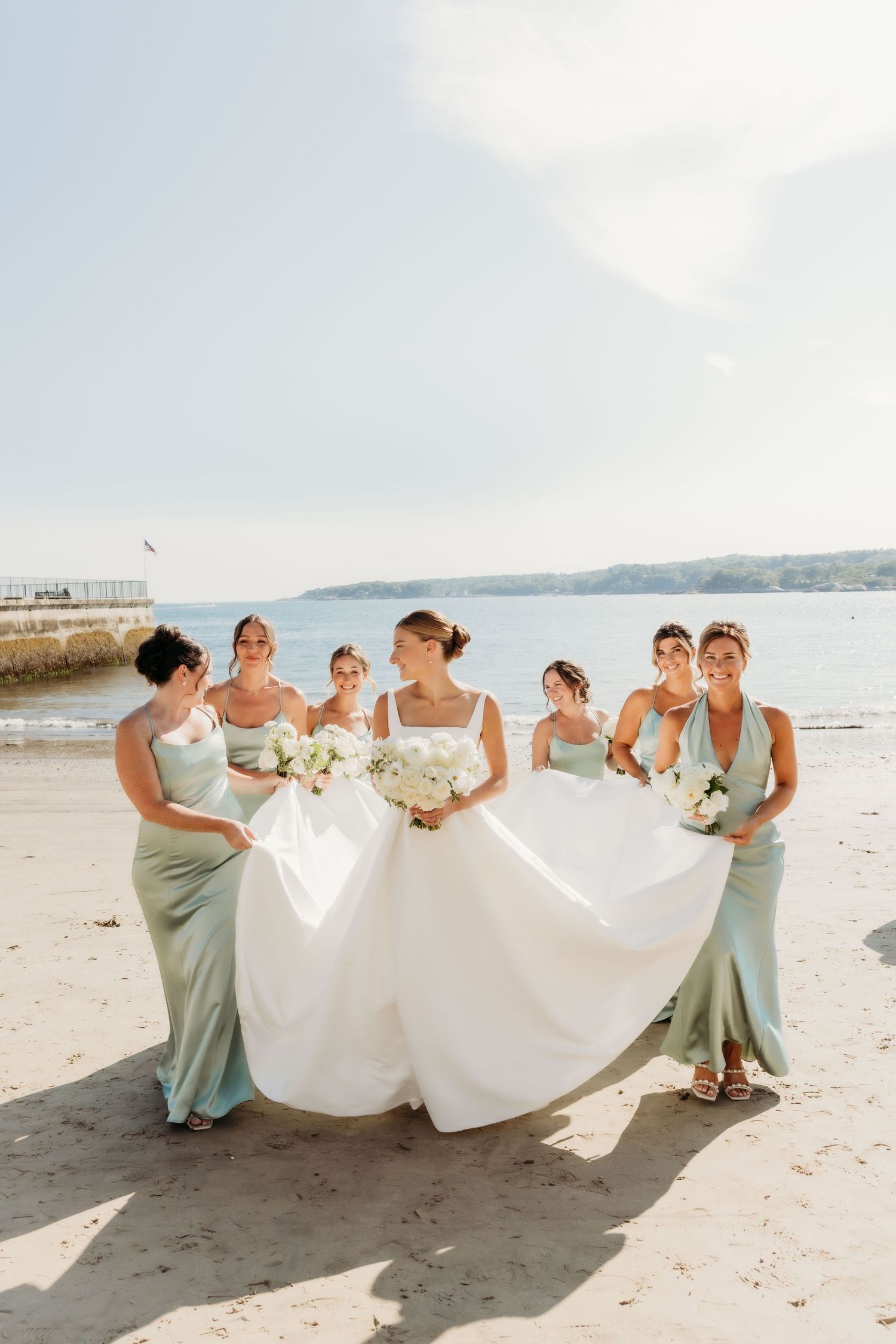 Bridesmaids in mint green dresses playfully holding up the bride's wedding dress train on a sunny beach.