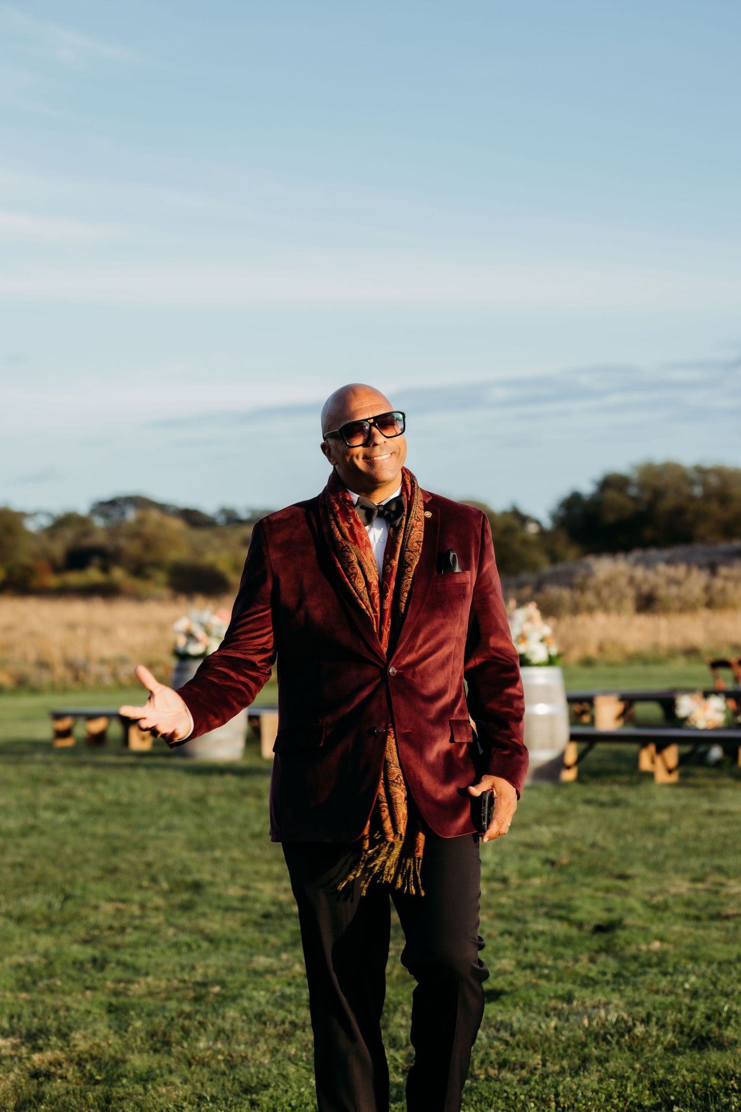 Someone wearing a burgundy blazer and sunglasses gestures while standing in a sunny field with pumpkins visible in background.