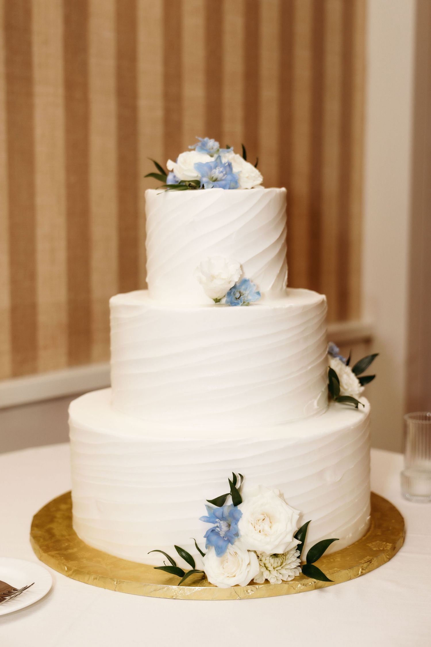 Elegant white wedding cake decorated with delicate blue hydrangeas and green leaves on a gold base.