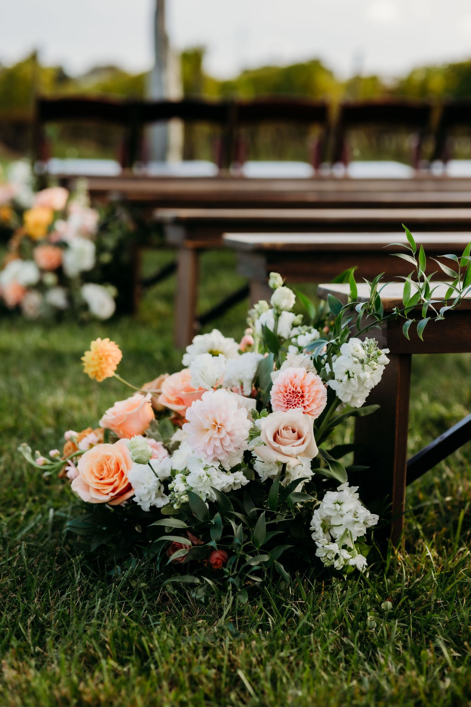 Beautiful floral arrangement with peach roses and white flowers decorates wooden chairs on grass at an outdoor wedding ceremony.
