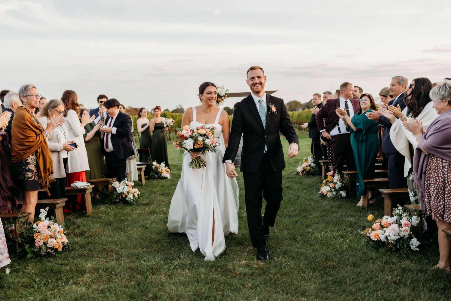 Wedding guests gather on a grassy lawn during an outdoor ceremony as the sun sets in the background.