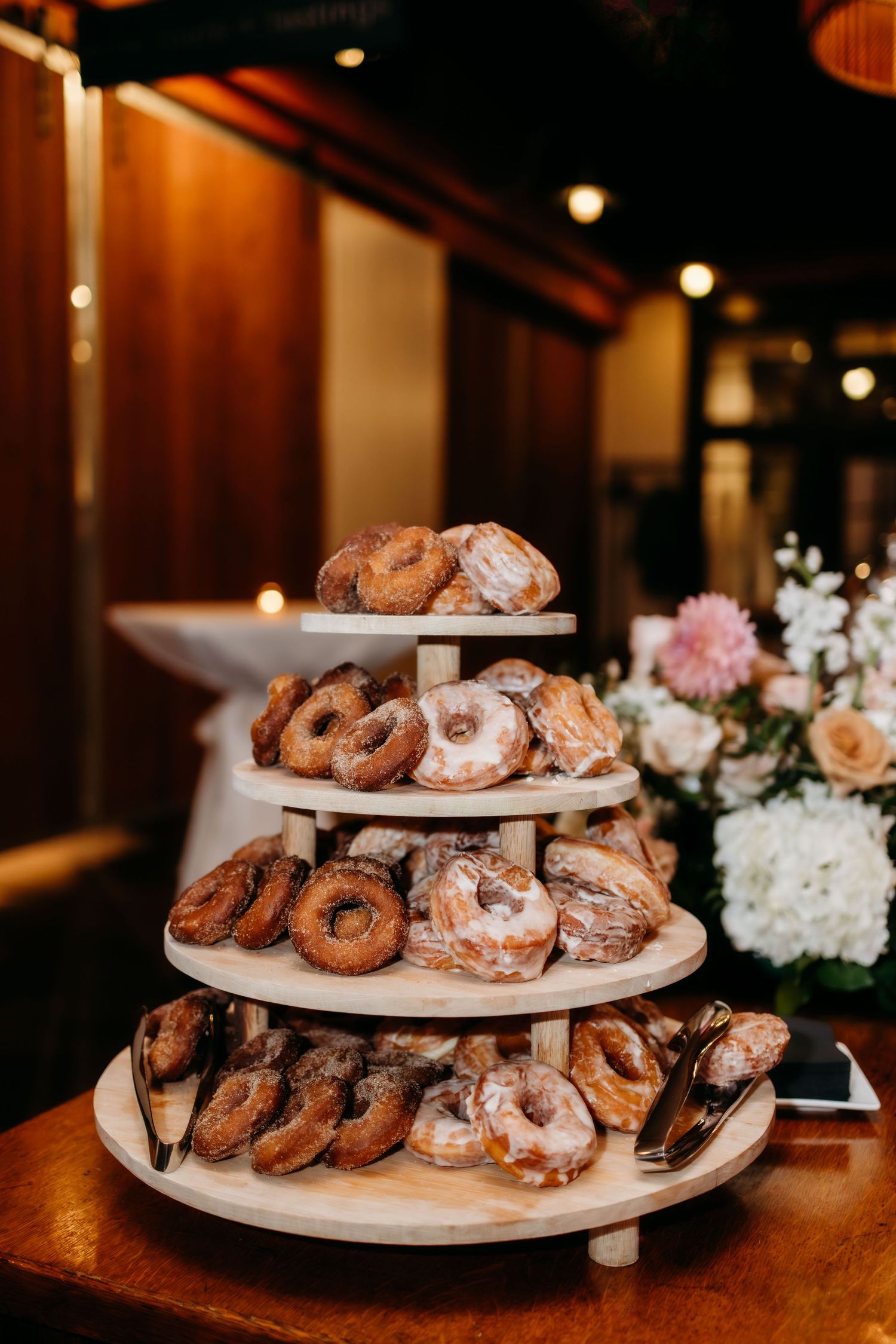 Tiered display of fresh donuts and pastries arranged on white plates at a rustic wooden table with soft lighting.