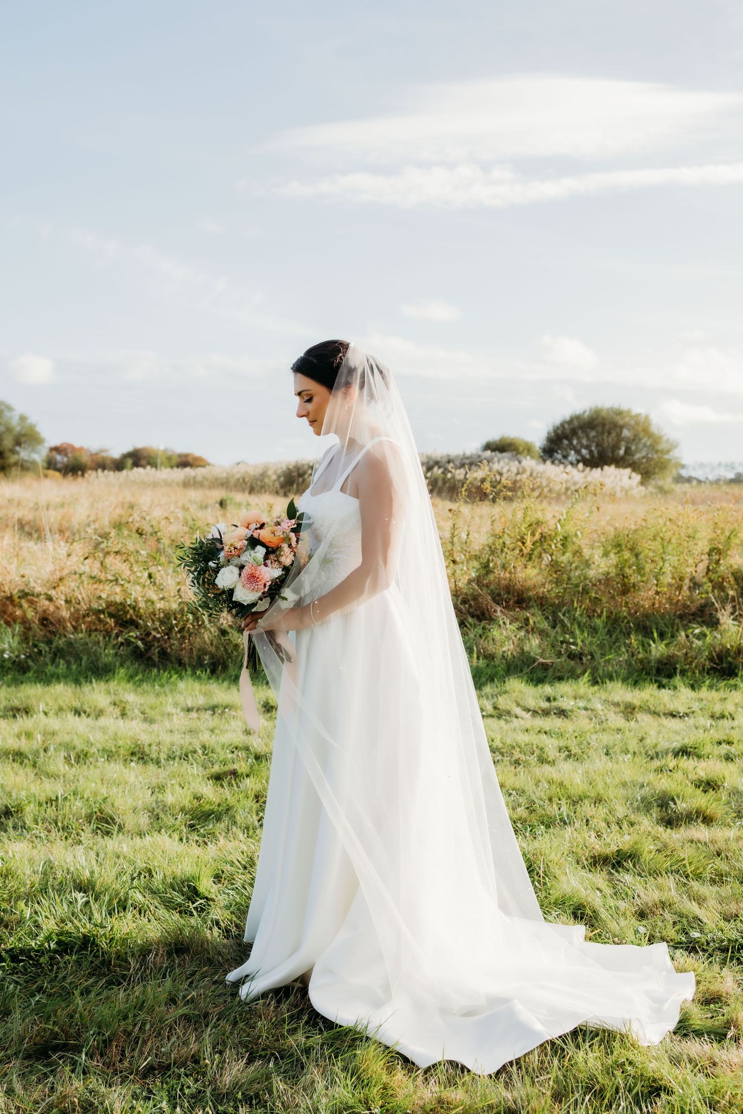 A bride in a flowing white wedding dress and veil stands in a sunlit meadow holding a pink and white bouquet.