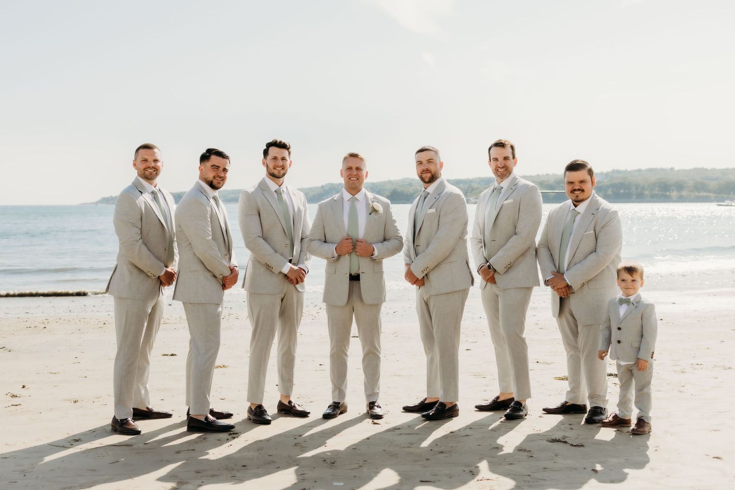A group of groomsmen in light gray suits stand together on a sunny beach with a young ring bearer.