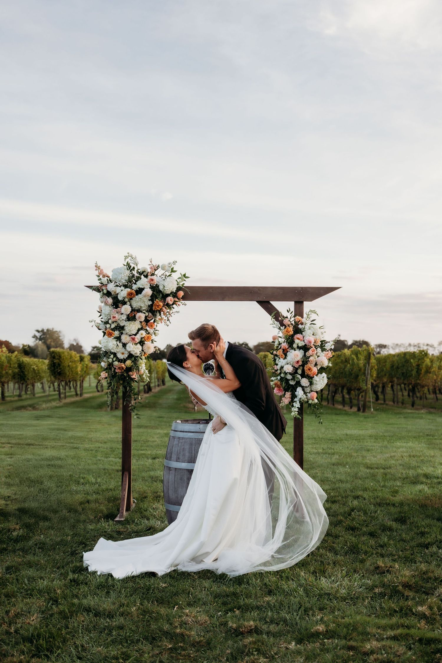 A romantic wedding couple shares a dip kiss under a wooden arch decorated with flowers in a scenic vineyard setting at sunset.