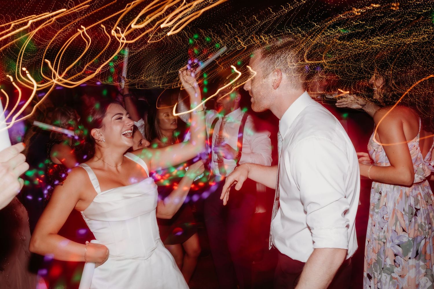 People dancing energetically at a wedding reception with light trails and colorful lighting creating a festive atmosphere.