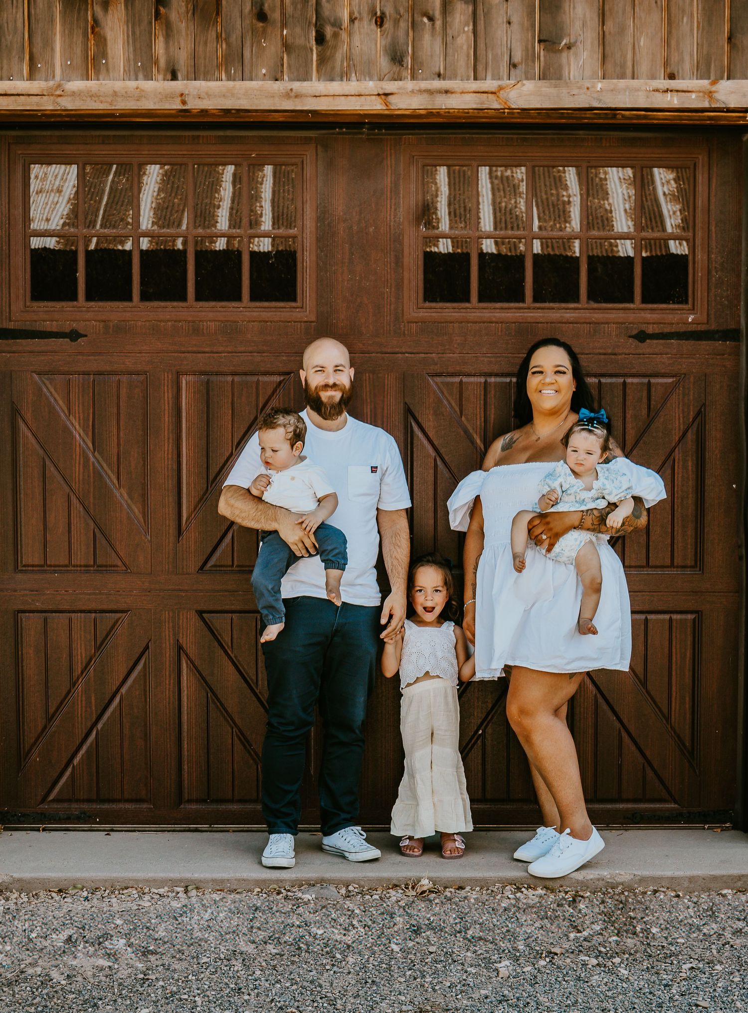 Happy family of five posing in white outfits before rustic wooden barn doors, parents holding two babies with toddler in middle.