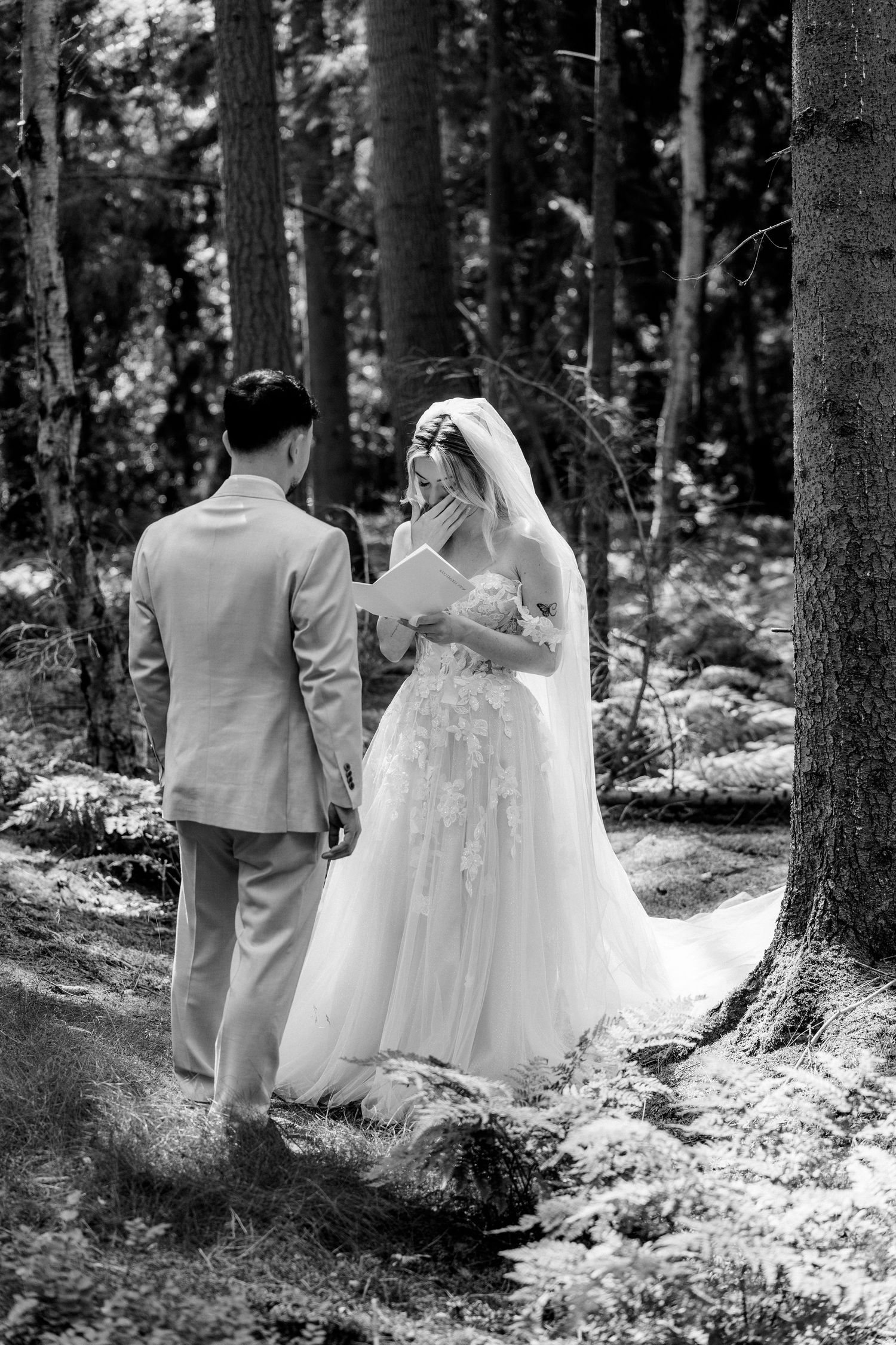 Black and white photo of bride and groom exchanging vows in a forest, bride emotional covering mouth.