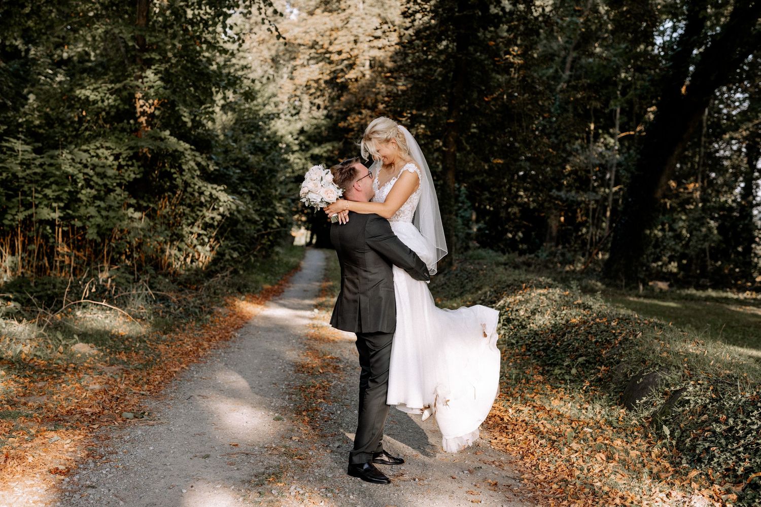 Newlywed couple embracing on a sunlit forest path covered in autumn leaves.