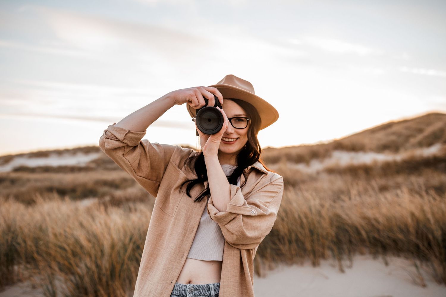 A photographer wearing a brown hat and beige coat takes pictures in a grassy coastal landscape at sunset.