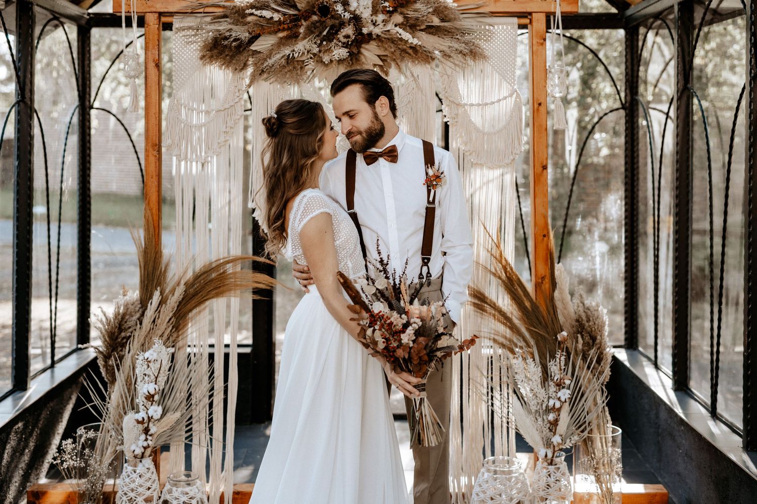 Wedding ceremony setup with macrame arch and dried flower arrangements in glass greenhouse.