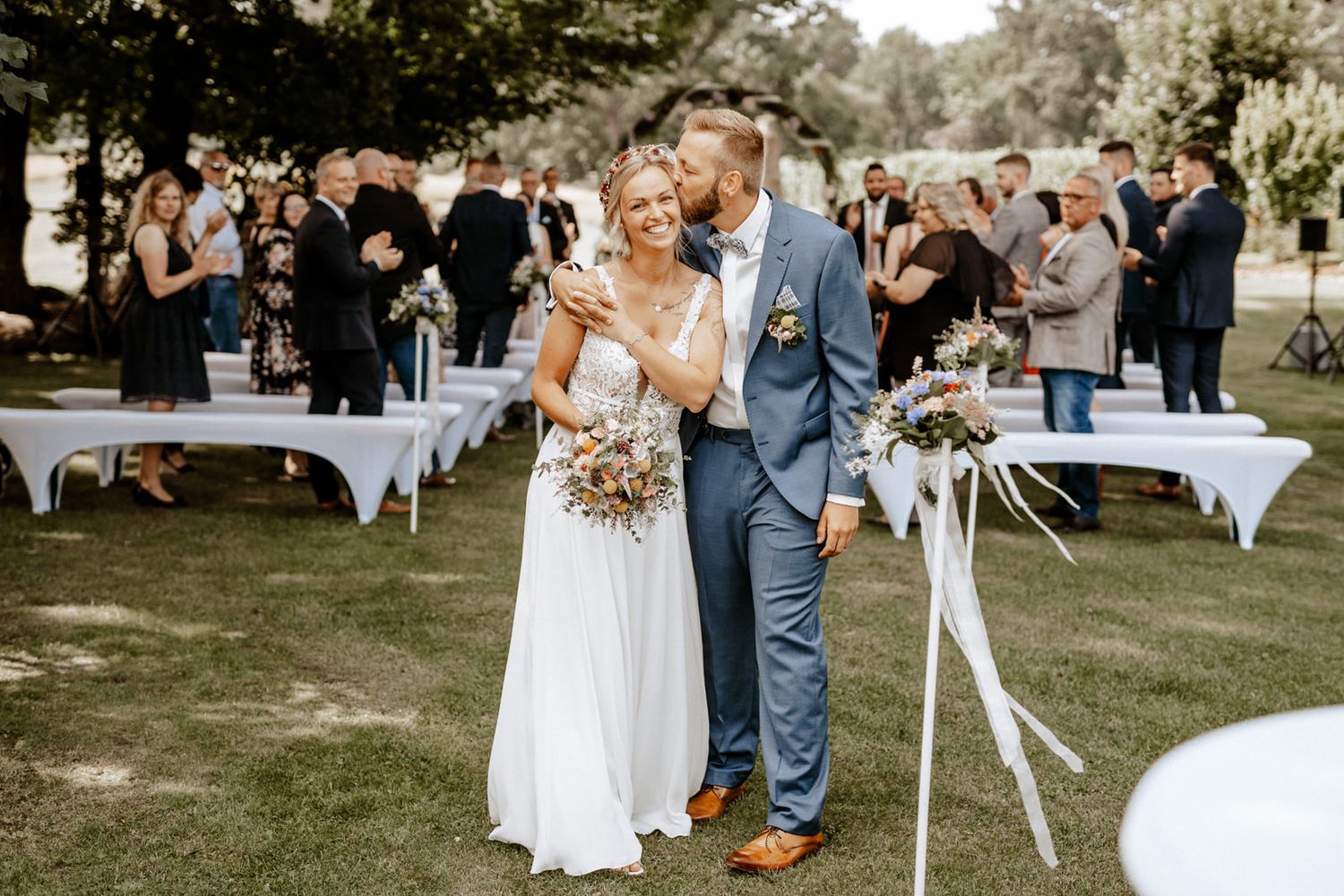 Garden wedding ceremony with guests and white tables in background.