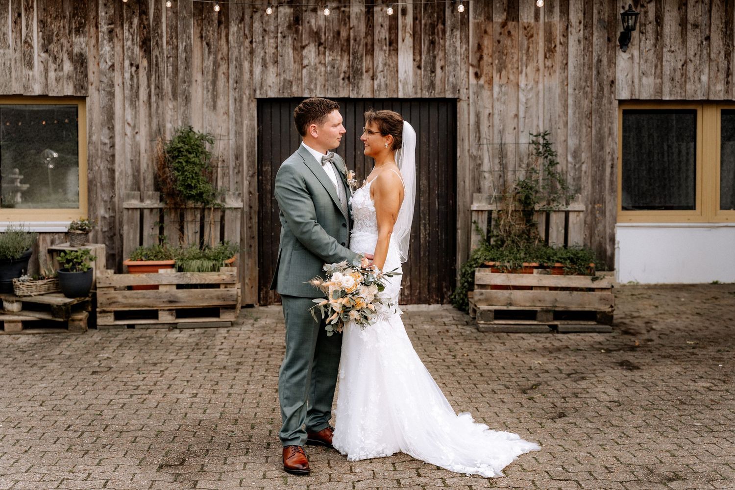 Bride and groom share a romantic moment in front of a rustic barn, bride holding a boho bouquet.