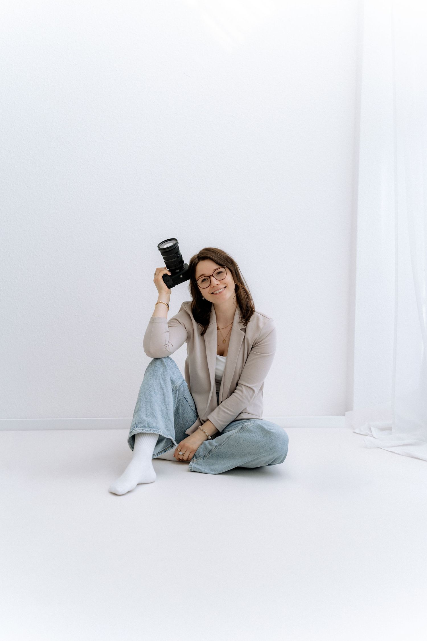 Professional photographer in a gray suit poses with camera equipment in a minimalist white studio setting.