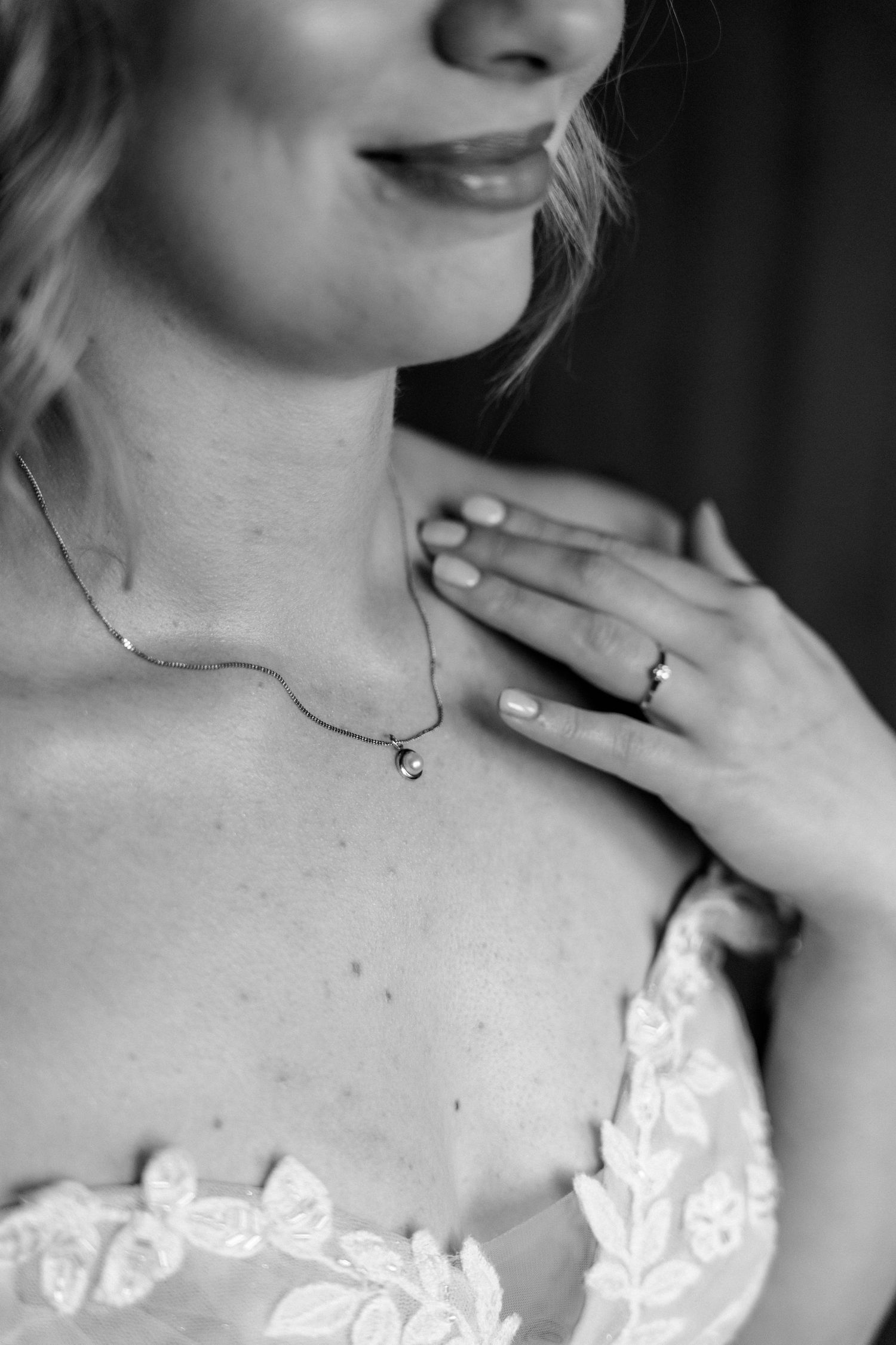Black and white photo of a bride wearing a delicate necklace and ring, hand placed on chest, in lace wedding dress.