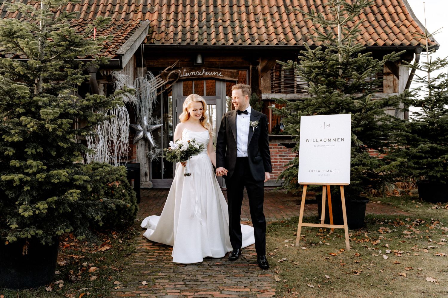Bride and groom walking hand in hand outside a rustic barn wedding venue surrounded by evergreen trees in winter.