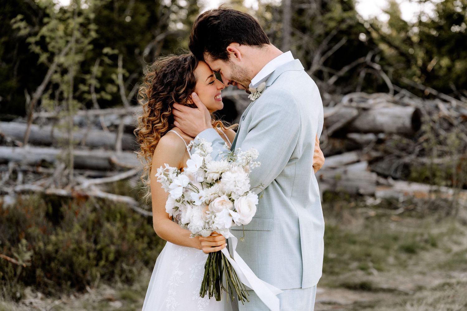 Newlywed couple embraces while holding white floral bouquet on rustic beach with driftwood in background.