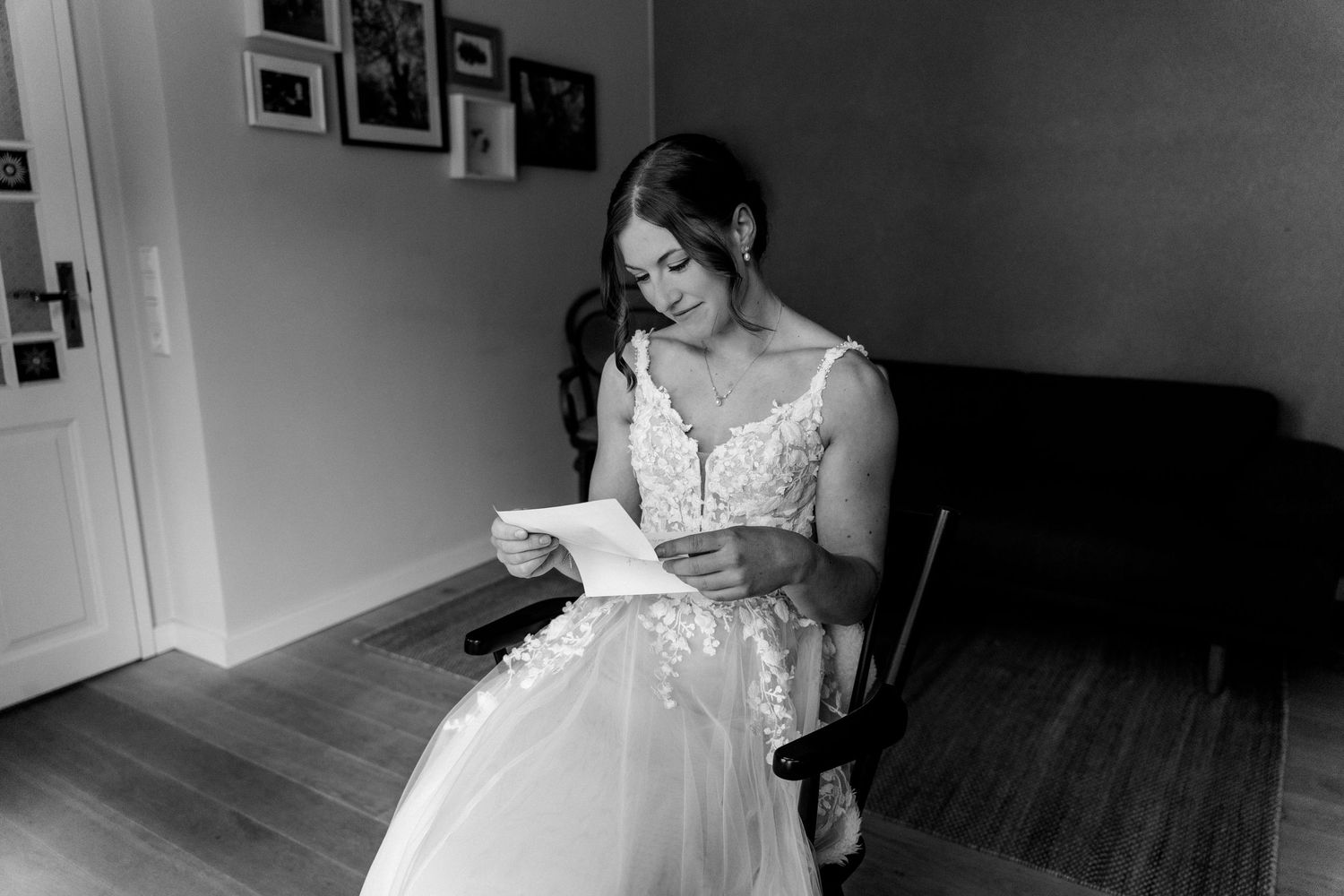 Bride in a lace wedding gown reads a heartfelt letter while seated, captured in elegant black and white photography.