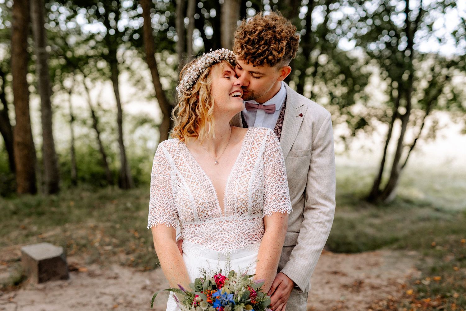 A couple shares a romantic moment outdoors, bride in lace gown holding wildflower bouquet, groom in beige suit.