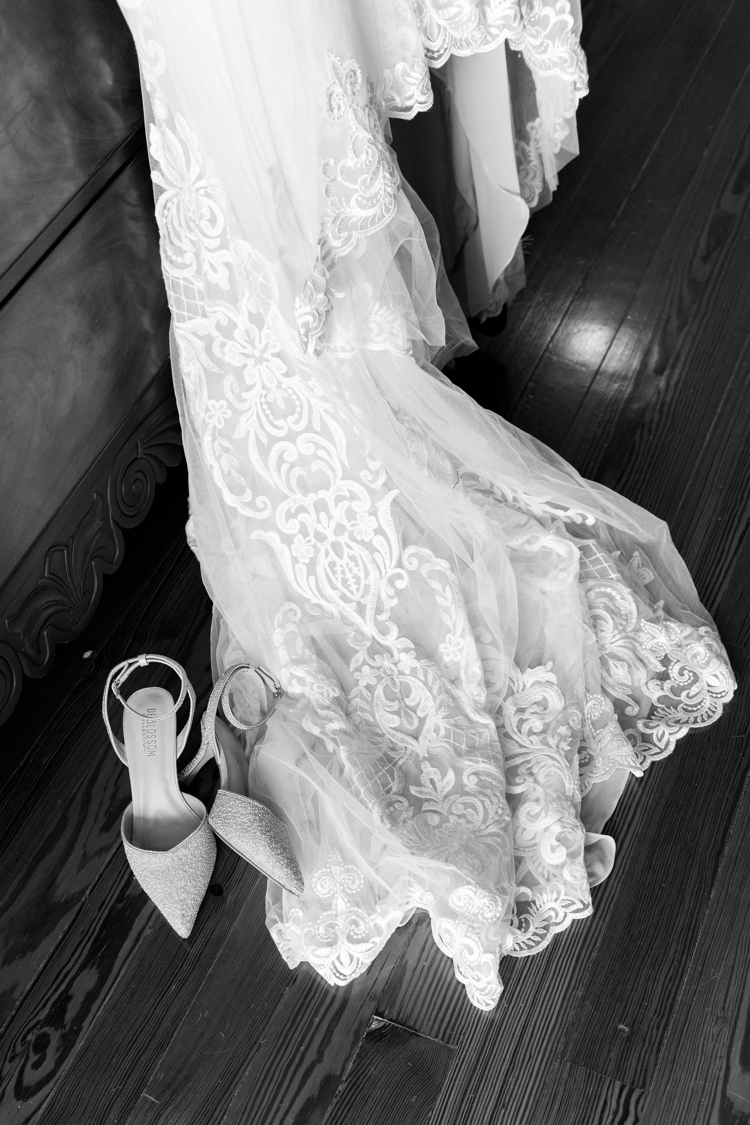 Black and white photo of a lace wedding dress hem and elegant high heel shoe on a dark wooden floor.