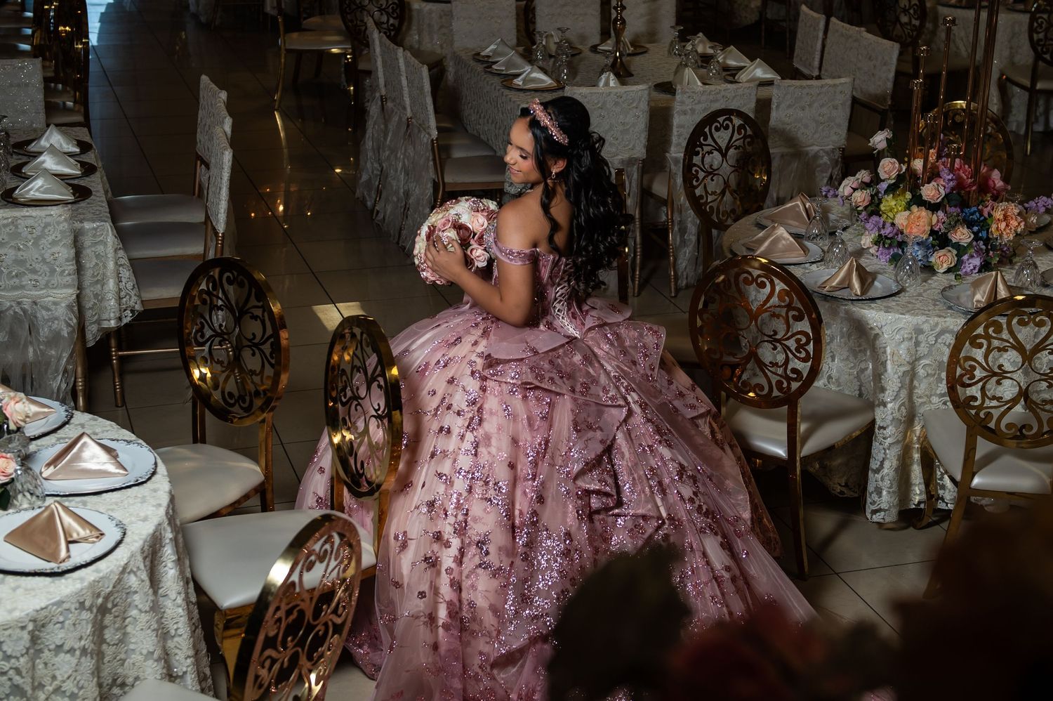Young woman in a pink floral ball gown at an elegant quinceañera banquet hall with gold chairs.