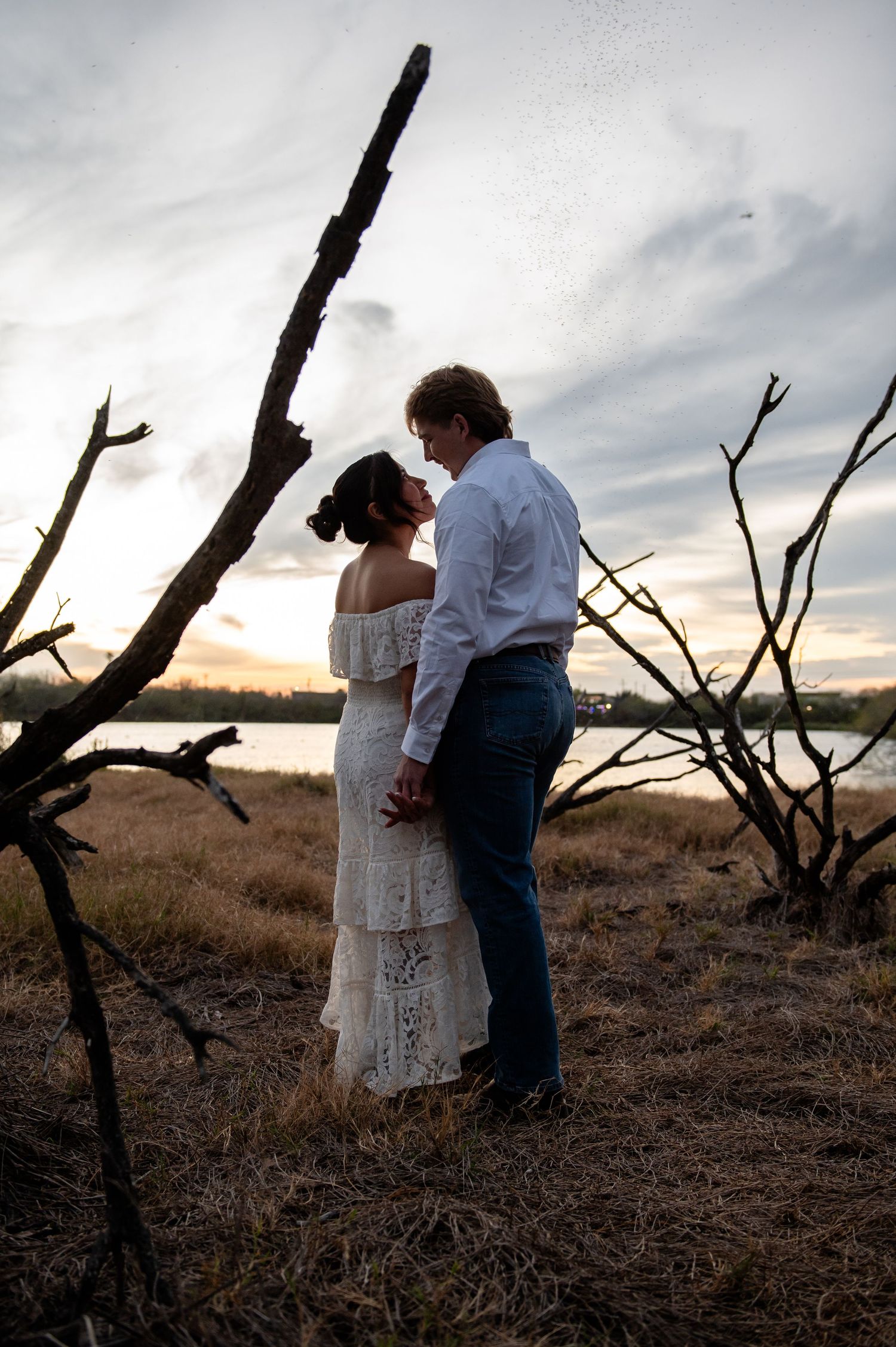 Couple sharing a romantic moment at sunset near a lake, woman in white lace dress and man in blue jeans embracing outdoors.