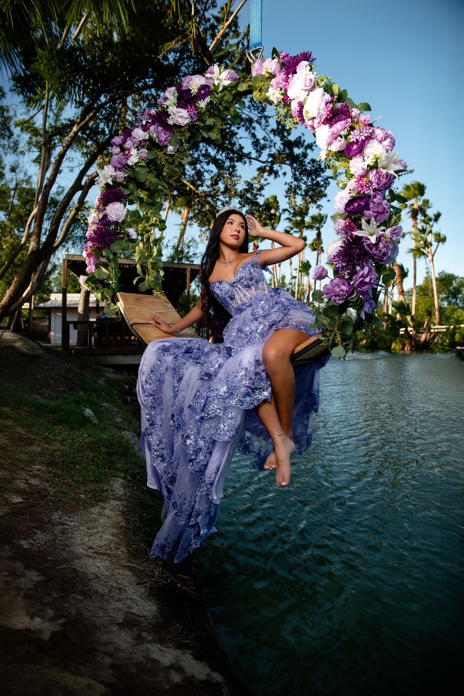 Young woman in lavender gown sits on floral arch swing over water surrounded by tropical trees.