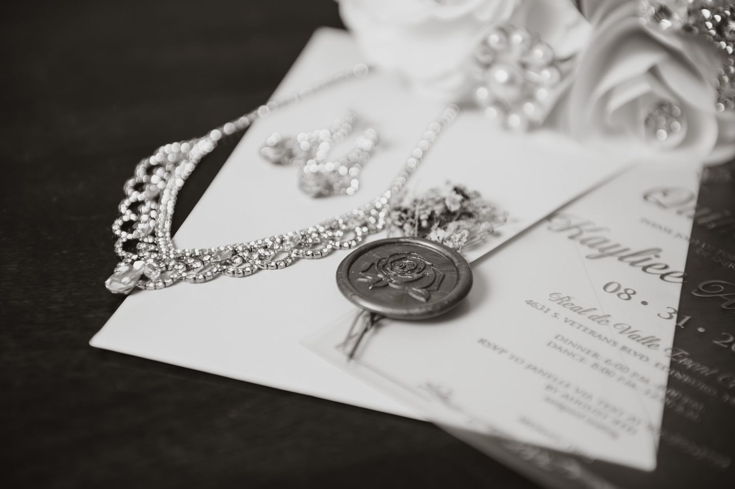 Elegant wedding flat lay with wax seal invitation, crystal tiara, diamond necklace, and white roses in black and white.
