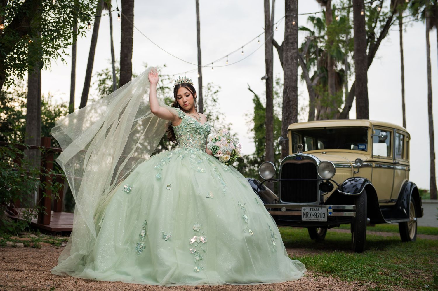 Young woman in mint green quinceañera ball gown poses with vintage yellow car amid tall palm trees.
