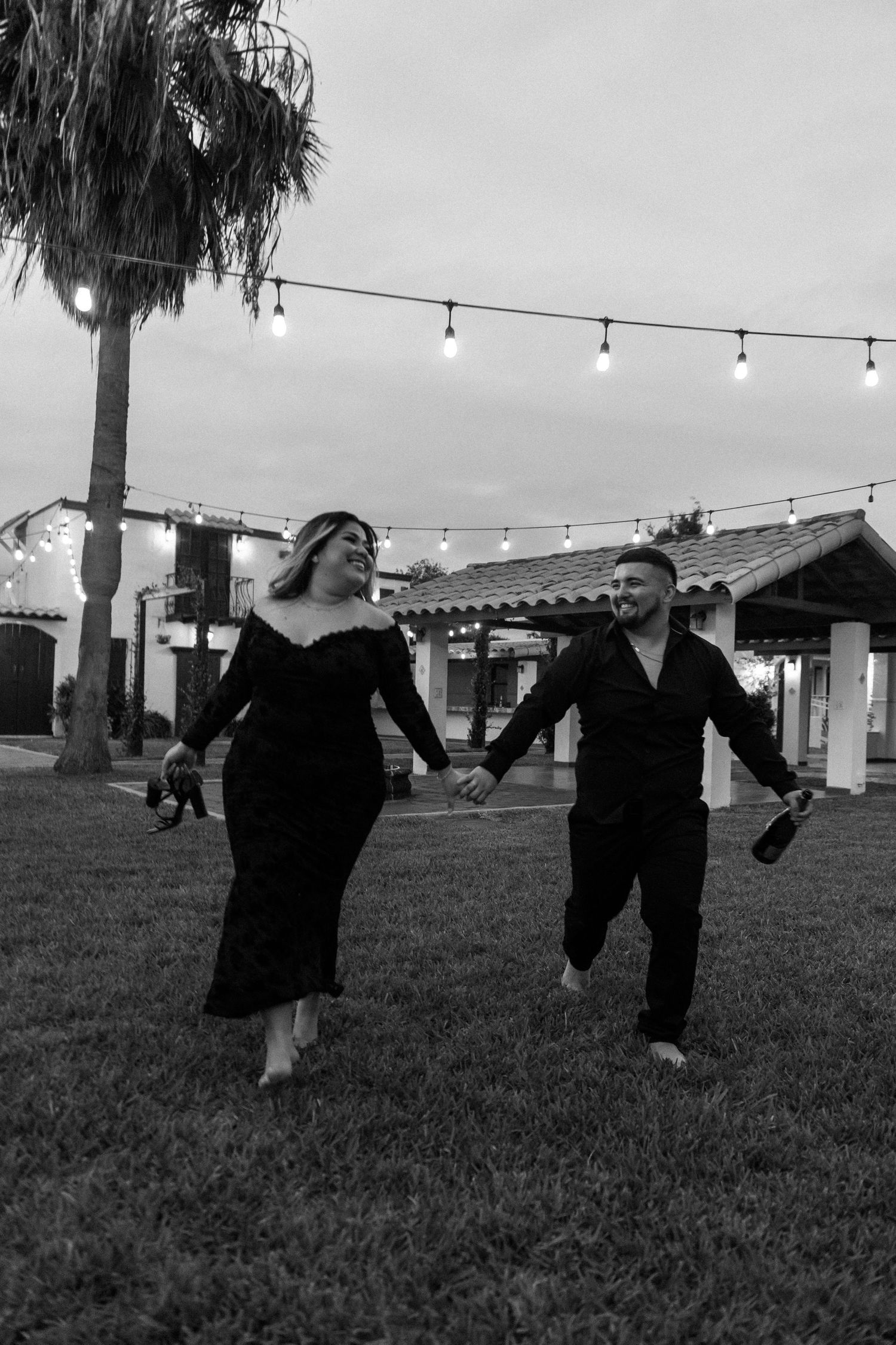 Black and white photo of a joyful couple holding hands and running on grass under string lights at dusk.