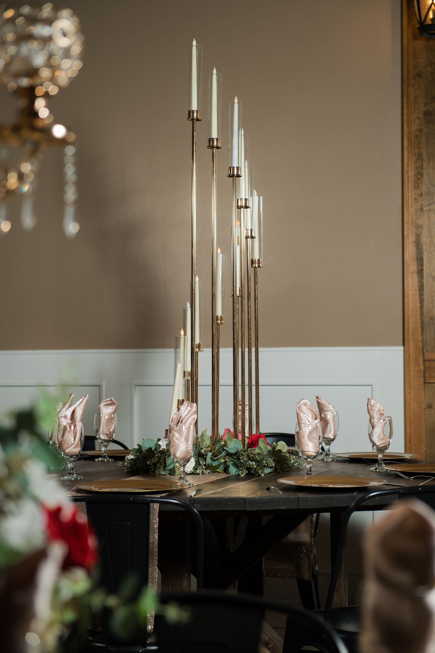 Elegant wedding table with tall gold candelabras, white taper candles, greenery centerpiece, and blush napkins on dark plates.