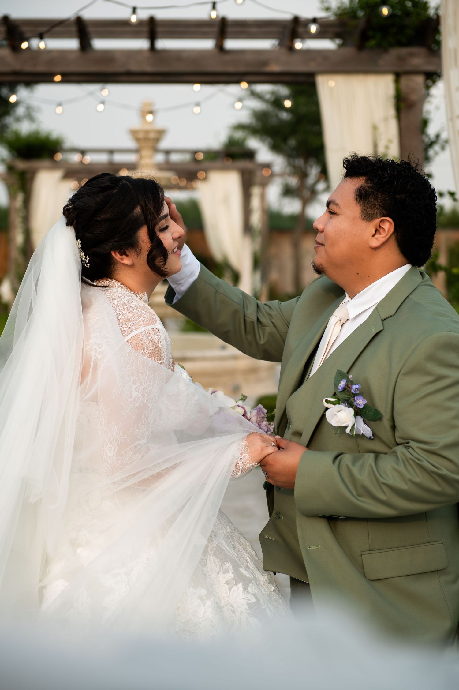 Groom in sage green suit tenderly touches bride's face during outdoor wedding ceremony with string lights.