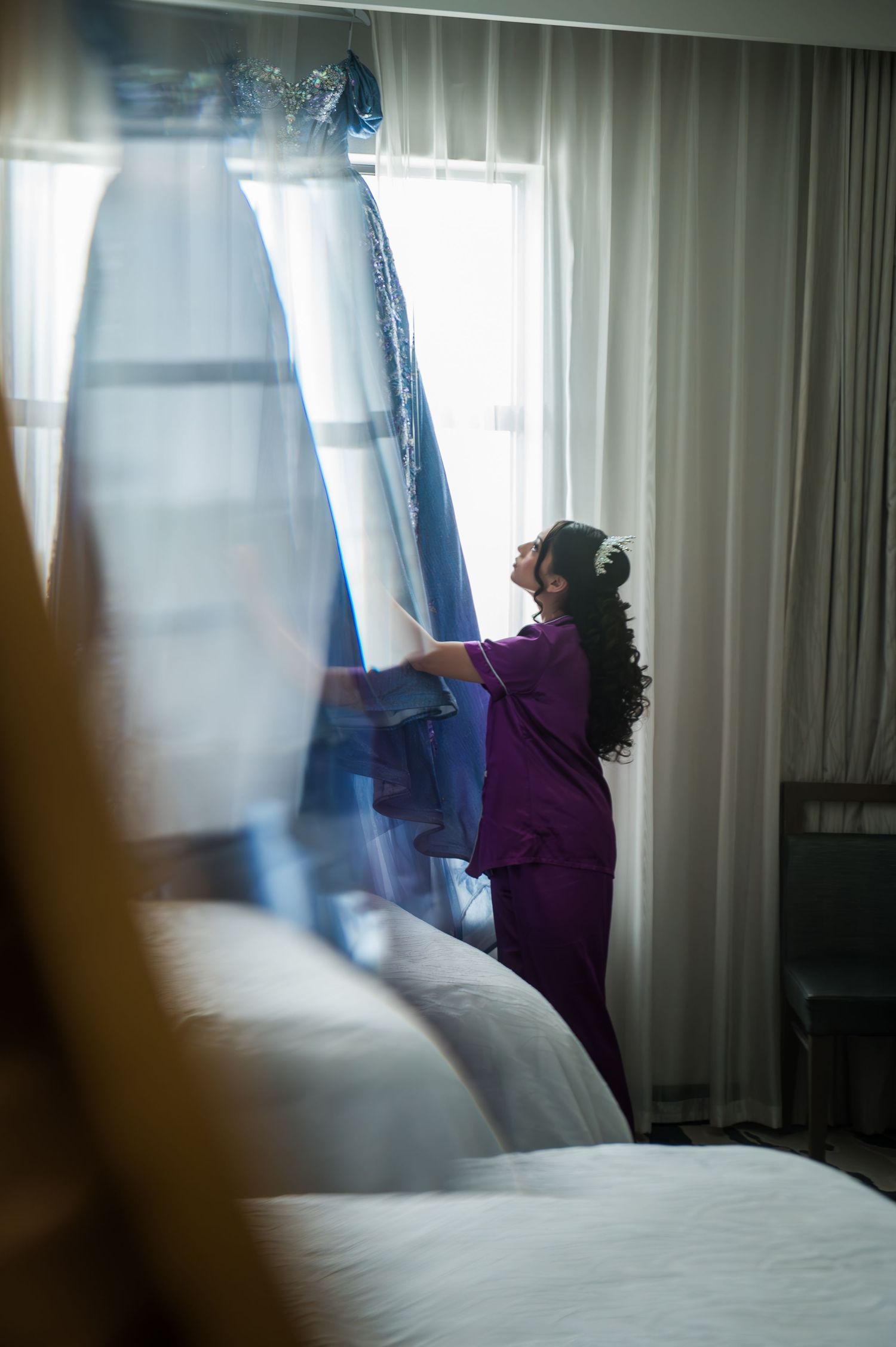 A woman in purple scrubs steams a blue wedding dress hanging by a bright hotel room window.