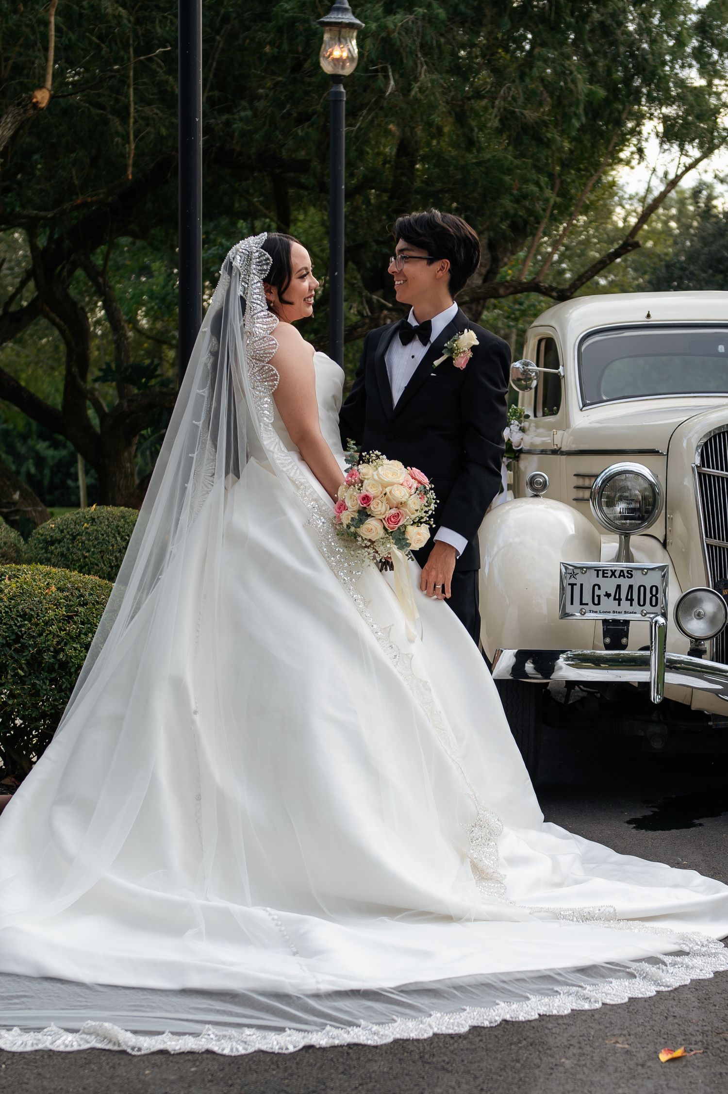 Bride in white gown with veil and groom in tuxedo pose beside a vintage Texas car surrounded by lush greenery.