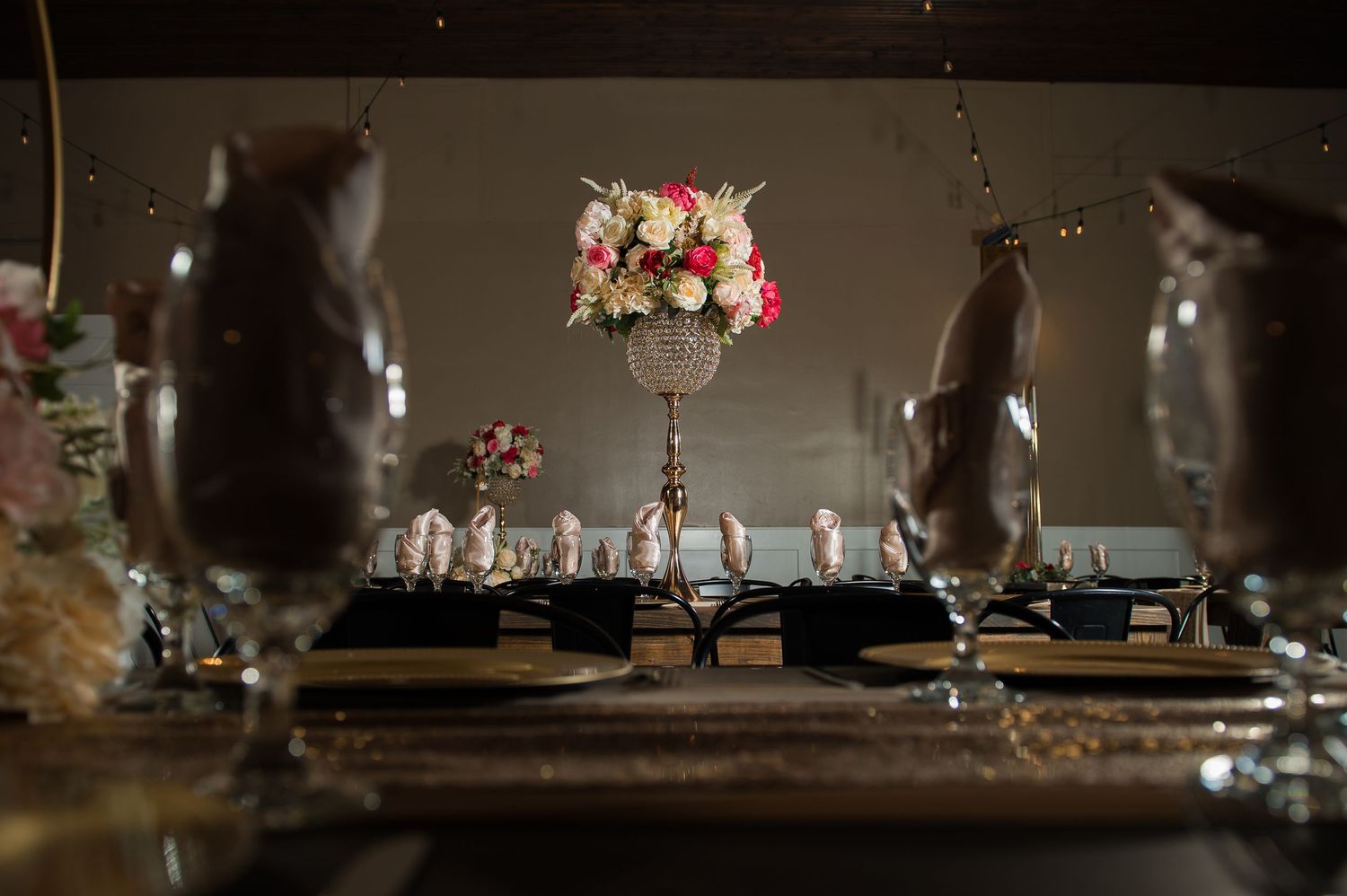 Elegant wedding reception table with tall floral centerpiece of pink and white roses, gold accents, and crystal glassware.