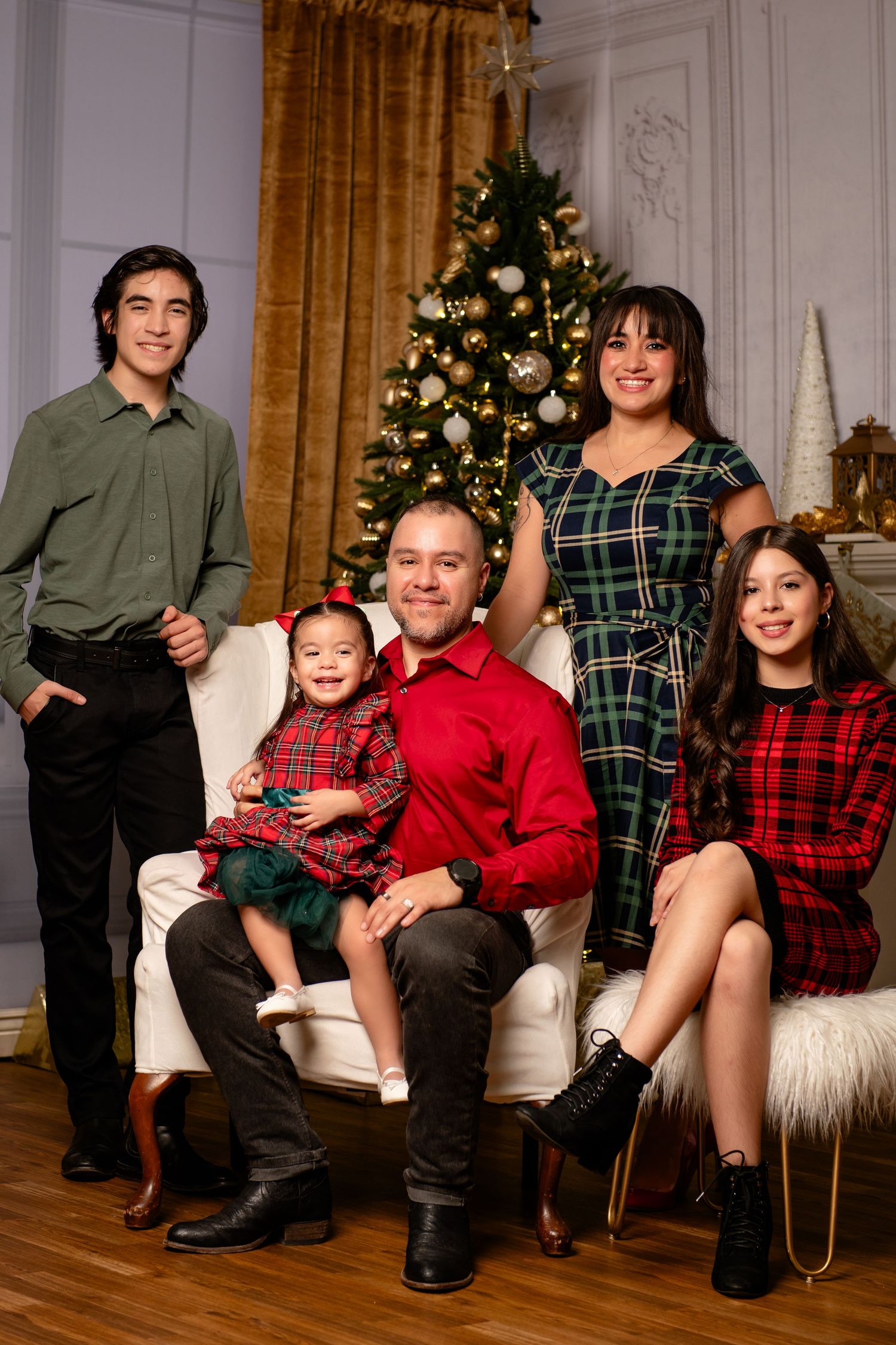 Family of five in matching plaid holiday outfits posing around a decorated Christmas tree in an elegant room with gold curtains.
