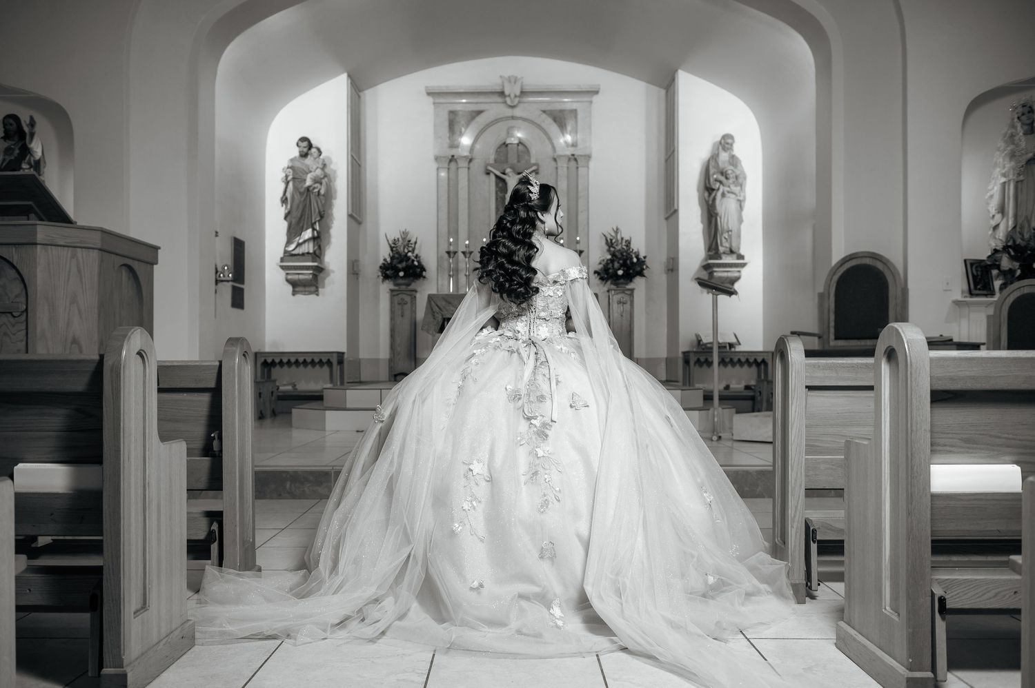 Black and white photo of a bride in a ballgown standing at a church altar, viewed from behind.