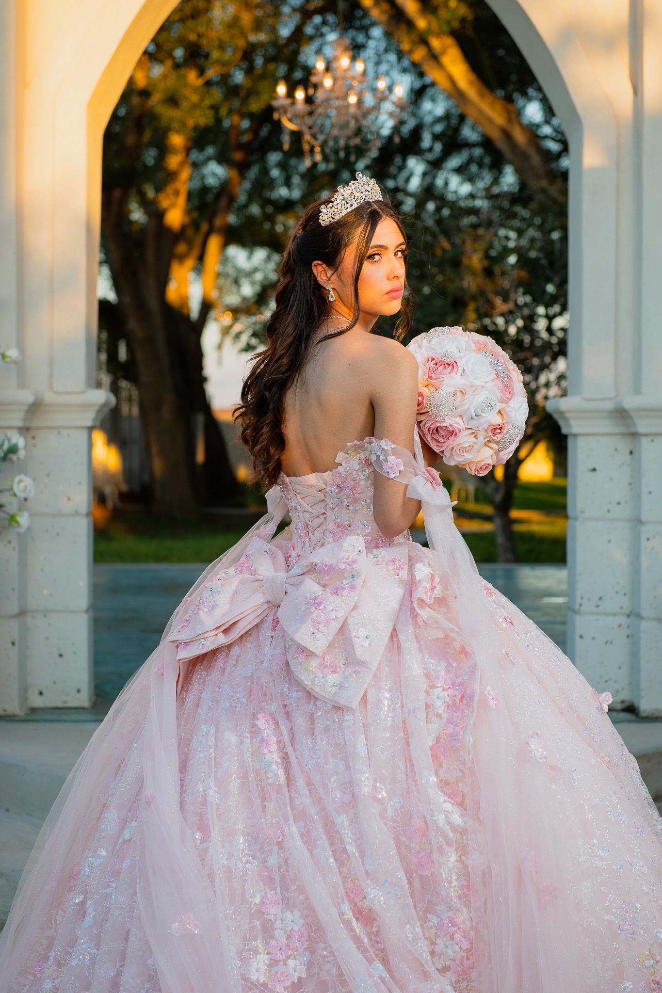 Young woman in pink ball gown with floral details, tiara, and bouquet poses under elegant arch at golden hour.
