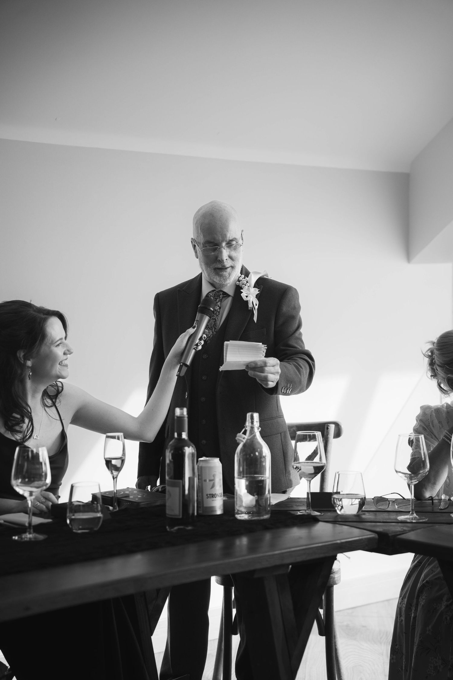 Bald man in suit delivers wedding speech while woman holds microphone at elegant reception table.
