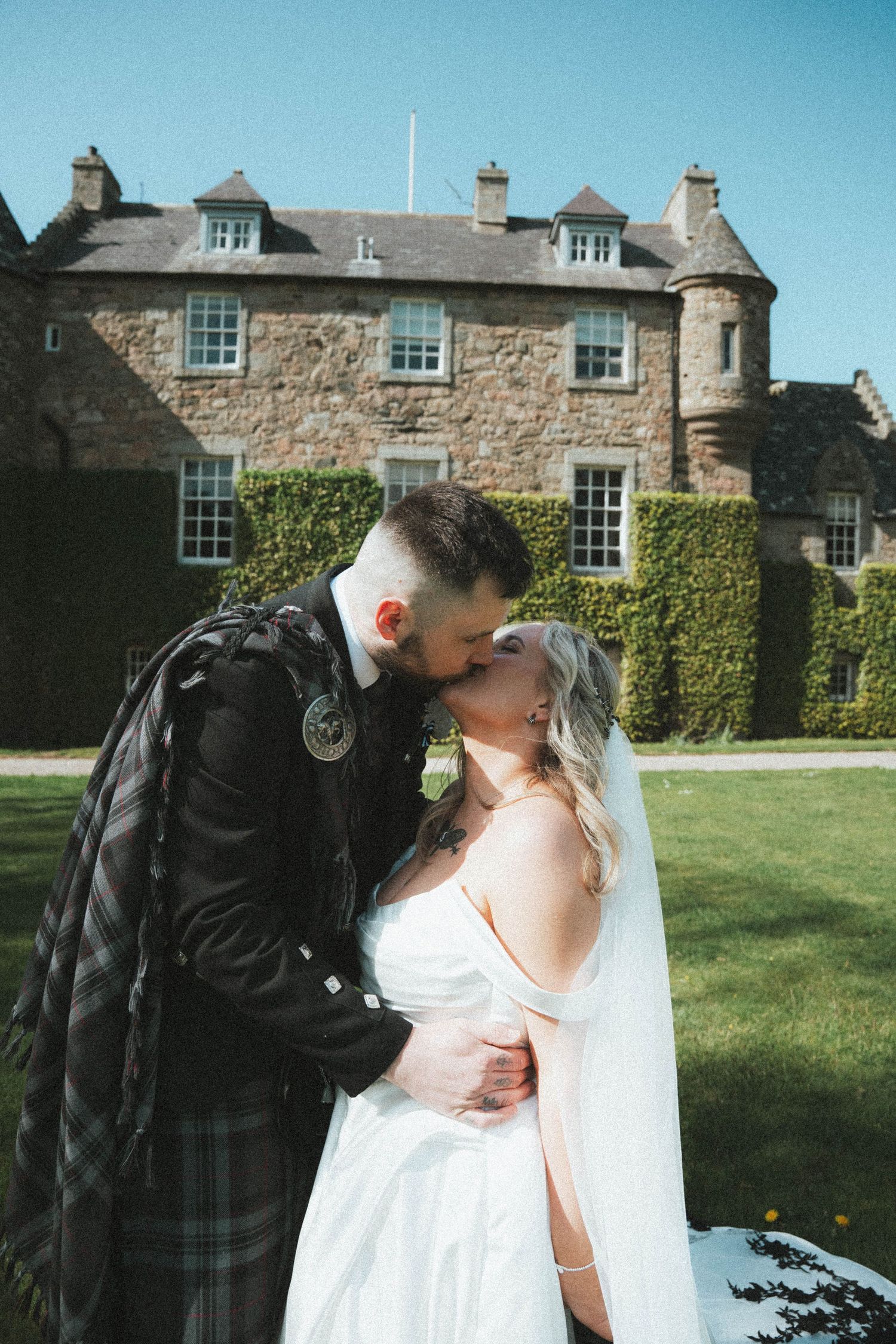 Bride and groom share a kiss on the lawn in front of a grand Scottish stone castle on a sunny day.