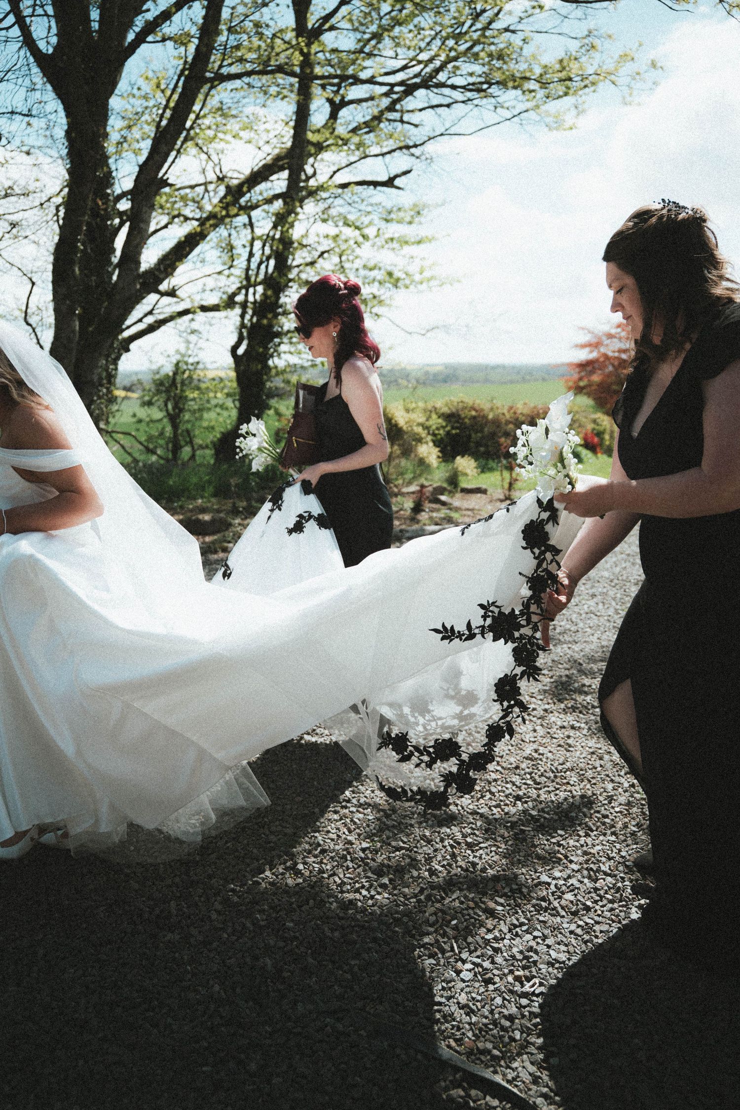 Bridesmaids in black dresses help carry the bride's white gown train on a sunny gravel path outdoors.
