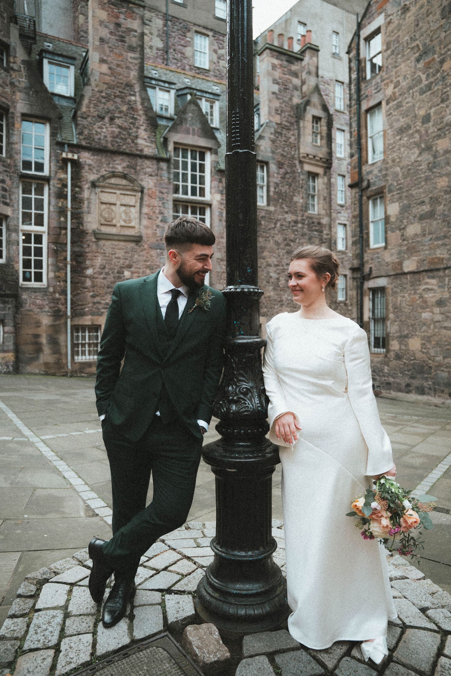A couple poses by a vintage lamppost in front of historic stone buildings.