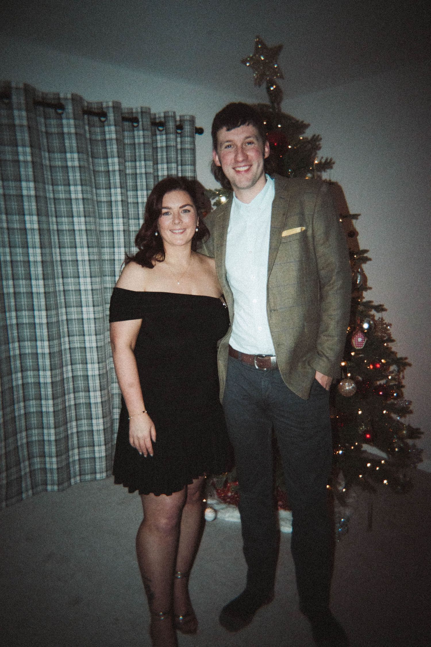 A couple poses in formal attire in front of a decorated Christmas tree.