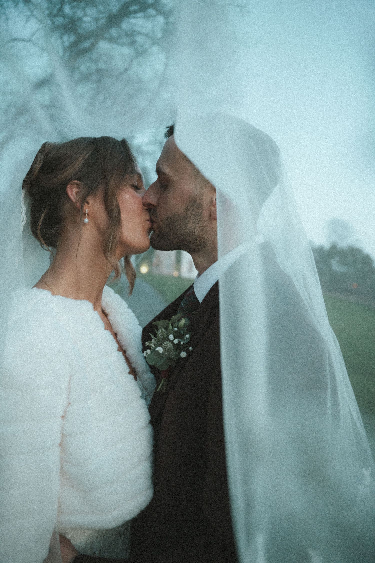Romantic wedding couple shares an intimate kiss under a flowing white veil against a misty outdoor backdrop.