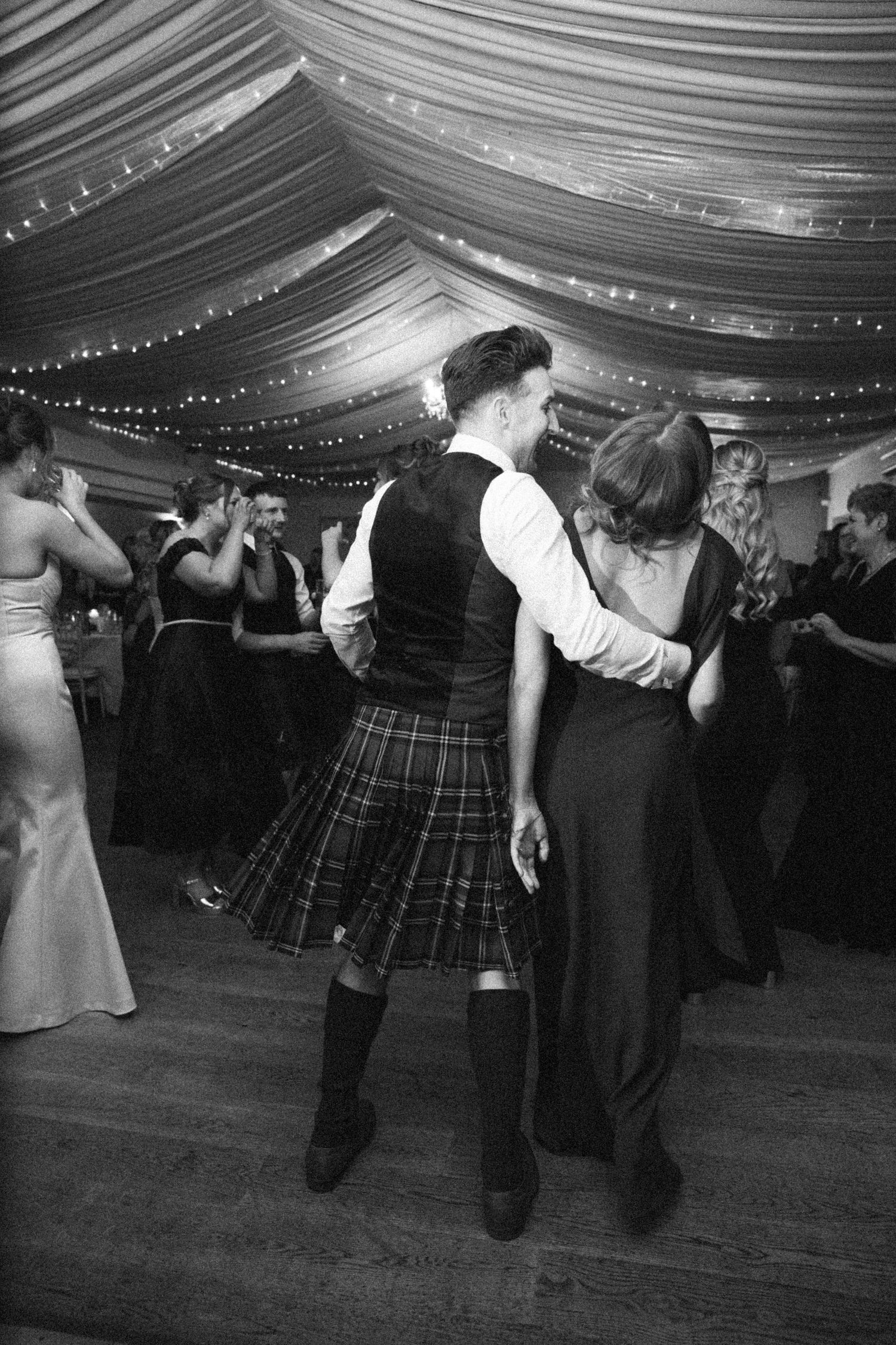 Wedding guests dancing in traditional Scottish kilts under elegant draped ceiling.