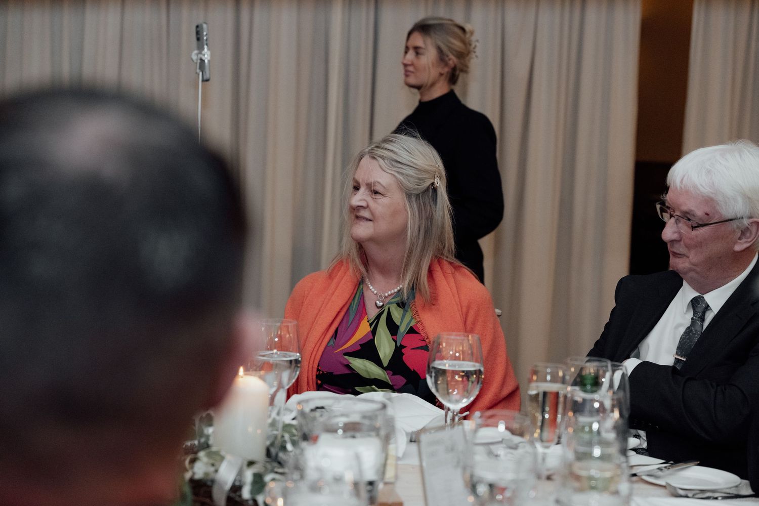 Wedding reception guests seated at decorated dinner tables with glassware and ambient lighting.