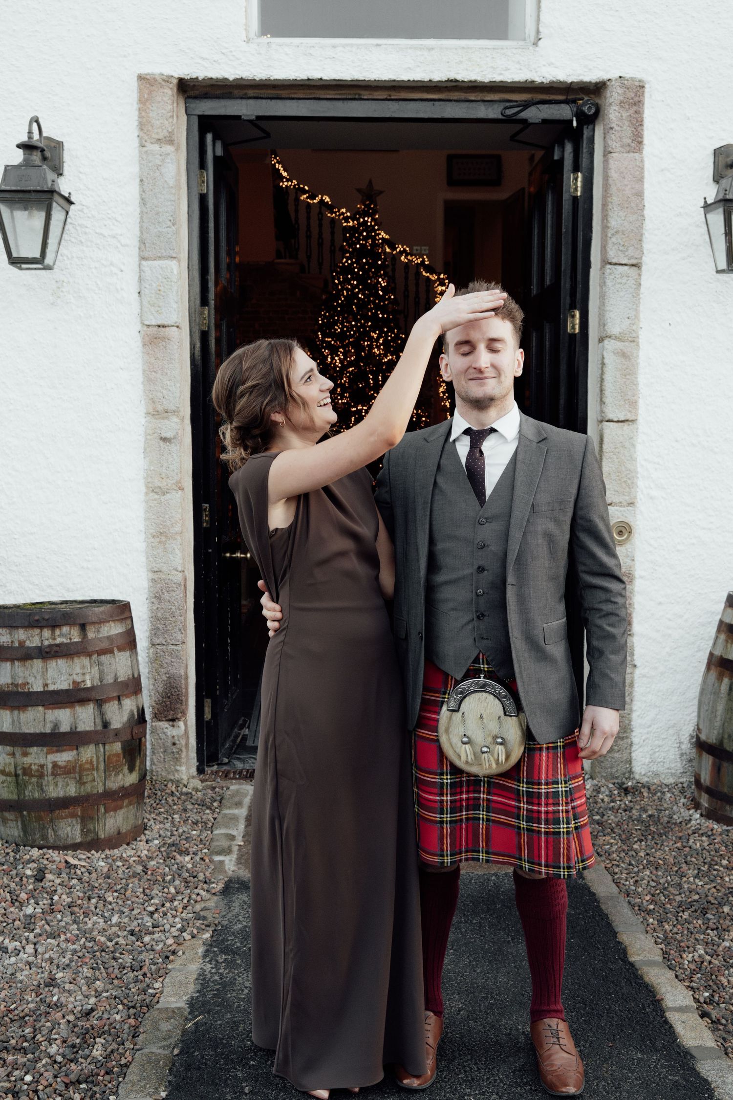 A couple poses playfully in front of a doorway with fairy lights, the man wearing a traditional Scottish kilt and suit.