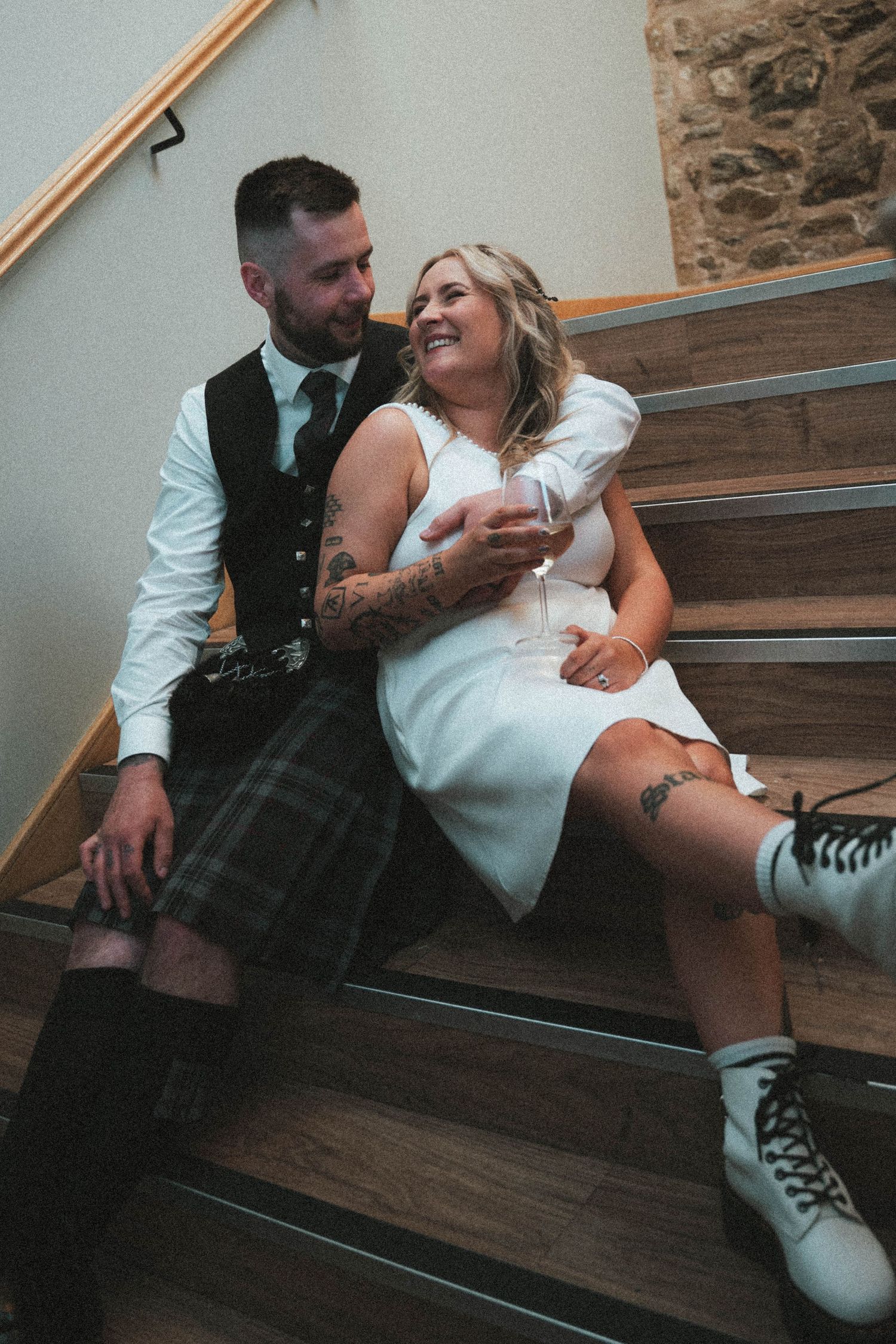 Tattooed bride in white dress and groom in tartan kilt sitting together on wooden stairs, smiling warmly.